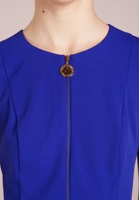 Blå klänning med rund halsringning, framzipper med en guldfärgad dekorativ dragkedja och en slät tygtextur.