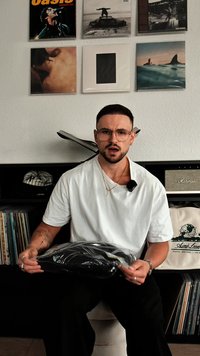 Un capo d'abbigliamento nero in un imballaggio trasparente tenuto da una persona che indossa una t-shirt bianca, seduta di fronte a un'esposizione e stoccaggio di dischi.