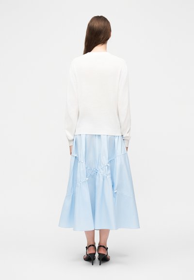 Γυναίκα όρθια με ορατή την πλάτη, φοράει λευκή μπλούζα με μακριά μανίκια, ανοιχτό μπλε midi φούστα με ρυτιδωμένες λεπτομέρειες και μαύρα πέδιλα με ψηλά τακούνια.