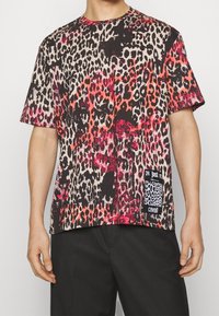 Man som bär en t-shirt med kort ärm i leopardmönster med röda, svarta och beige toner samt en liten svartvit patch med varumärkeslogotyp på framsidan.