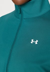 Tuurkleurig jack met een hoge kraag en een volledige rits. Gemaakt van gladde, rekbare stof. Heeft een wit logo aan de linkerkant.