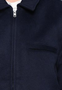 Tumši zila rāvējslēdzēja jaka no mīksta auduma ar klasisku apkakli un krūšu kabatu ar gludu, minimālistisku dizainu.