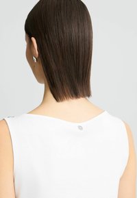 Top bianco senza maniche in tessuto morbido, con scollatura rotonda e un piccolo dettaglio di bottone argentato sul retro, indossato da un modello con capelli lisci.