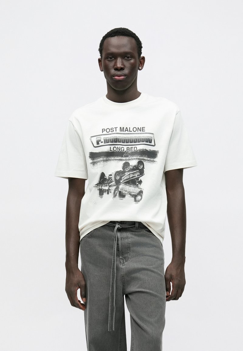 T-shirt de algodão branco com um design gráfico preto e os textos "Post Malone" e "LONG.BED". Combinado com calças cinzentas largas.