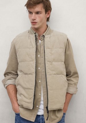 Young man wearing beige quilted vest over beige button-up shirt and white t-shirt, standing with hands in pockets against plain background.