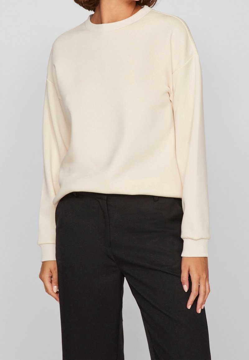 Femme portant un pull crème à manches longues et un pantalon noir taille haute sur un fond uni gris clair.