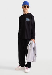 Schwarzes Langarm-T-Shirt mit blauer Grafik, lockere schwarze Hose, hält einen grauen Hoodie, blaue Strickmütze und weiße Sneakers mit pinken Akzenten.