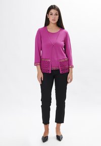 Fuchsia Cardigan mit Dreiviertelärmeln, versehen mit Spitzendetails und goldenen Akzenten, kombiniert mit schwarzen Hosen und spitzen Ballerinas.
