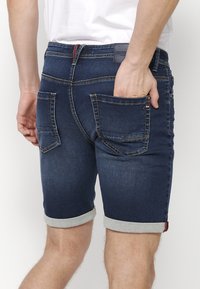 Homme portant un short en denim bleu foncé retroussé avec des poches arrière, une main placée dans la poche droite, chemise blanche visible au-dessus.
