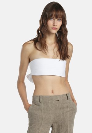 Femeie cu păr lung, ondulat, castaniu, purtând un top bandeau alb fără bretele și pantaloni bej din in, cu mâinile în buzunare.