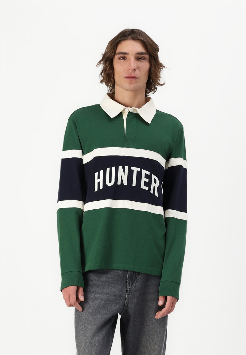 Grønn langermet polo skjorte med en hvit og marineblå horisontal stripe med teksten "HUNTER" i fete hvite bokstaver. Klassisk krage design.