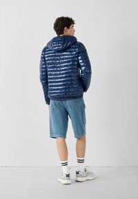 Marineblauwe pufferjas met een glanzende afwerking, horizontaal naaipatroon en een capuchon. Draagt met denim shorts en sneakers.