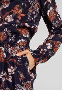 Hånd i lommen på navy beklædning med rødt og hvidt blomstermønster samt elastisk talje og manchetter.