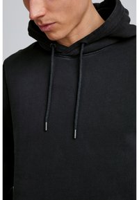 Melns kapuci svārkos ar auklām un metāliskām galiem. Gluda tekstūra, ikdienišķs dizains, ar ribinātiem piedurknēm un apakšmalu.