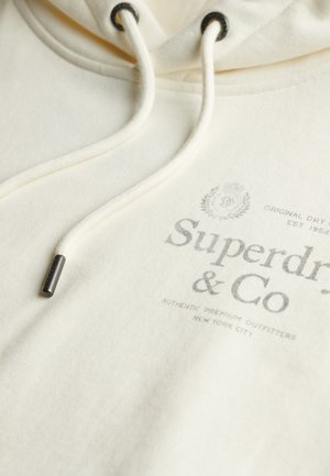 Sweat à capuche couleur crème avec capuche à cordon de serrage. Dispose d'un logo imprimé en gris et d'accents métalliques sur les cordons. Tissu doux et texturé.