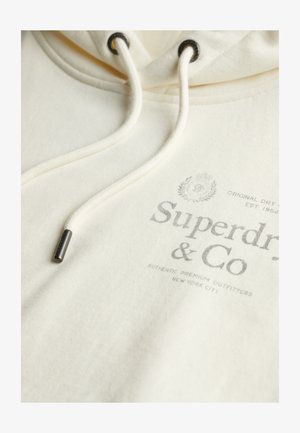 Sweat à capuche couleur crème avec capuche à cordon de serrage. Dispose d'un logo imprimé en gris et d'accents métalliques sur les cordons. Tissu doux et texturé.