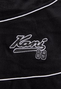 Material textil de culoare neagră cu o textură moale, având margini albe. Un logo brodat care spune "Kani 89" într-un font stilizat.