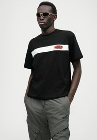 T-shirt de algodão preto com uma faixa horizontal branca e logo vermelho. Combinado com calças cargo cinzas e óculos de sol bege.