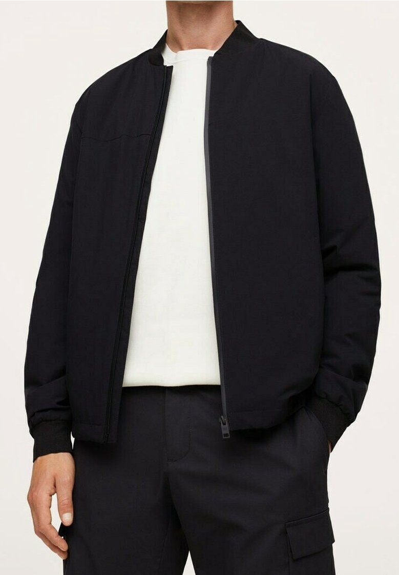 Veste bomber noire avec un col haut, fermeture éclair et poignets côtelés. Portée sur une chemise blanche, associée à un pantalon foncé. Matière lisse.