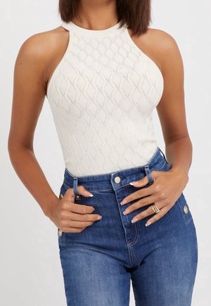 Kobieta ubrana w białą, bezrękawnikową, teksturowaną dzianinową bluzkę wsadzoną w wysokiej talii niebieskie dżinsy z detalami ze złotymi guzikami.