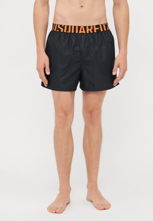 Torse et jambes masculins portant un short de bain noir avec une large ceinture élastique comportant des lettres orange vives, debout pieds nus sur un fond blanc.