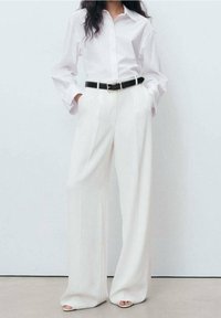 Chemise blanche oversize à manches longues, ceinture noire, et pantalon blanc à larges jambes taille haute ; tissu lisse avec des lignes épurées.