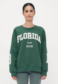 Zöld pulóver, rajta fehérrel nyomtatva "FLORIDA" és "MIAMI" felirat. Laza szabású, kerek nyakkivágású, hosszú ujjú, a mandzsettákon fehér részletekkel.