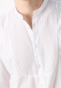 Camisa blanca de botones con un diseño de frente plisado, que presenta un cuello clásico y pequeños botones blancos a lo largo de la tapeta.