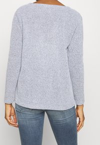 Osoba w teksturowanym jasnoszarym swetrze z długim rękawem i niebieskich dżinsach w średnim odcieniu, pokazana z tyłu.