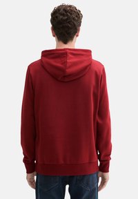 Felpa rossa in cotone con cappuccio regolabile, polsini e orlo a costine. Tessuto morbido, vestibilità comoda, design semplice, indossata sopra jeans blu.