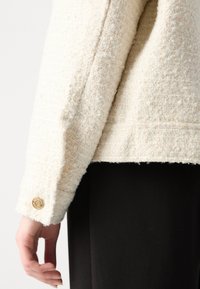Cremefarbener Blazer aus strukturiertem Stoff mit goldenen Knopfdetails an den Manschetten. Das Design ist strukturiert und hat eine taillierte Passform.