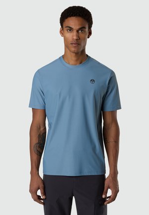 Uomo con capelli corti e ricci, che indossa una camicia azzurra a maniche corte e pantaloni scuri, in piedi davanti a uno sfondo semplice.