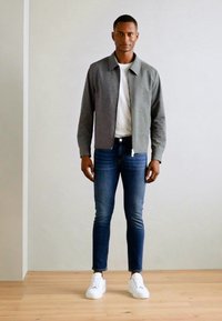 Siva zip-up jakna preko bijele majice, uparena s tamnoplavim trapkama i bijelim tenisicama. Minimalistički dizajn s glatkim teksturama.