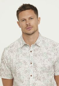 Chemise à manches courtes en tissu léger avec une base blanche, ornée d'un motif de feuilles vertes et brunes, et de boutons bruns.