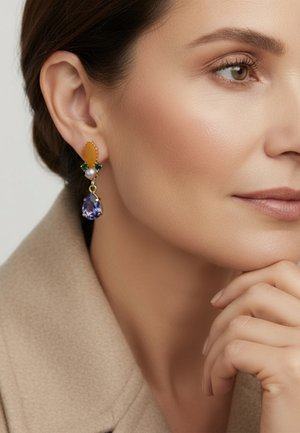 Vrouw in een beige jas die een multicolor druppelvormige oorbel met gele, groene, parel- en paarse edelstenen toont, hand rustend vlakbij de kin.