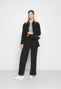 Black oversized shirt jacket over a grey top and black wide-leg trousers. Model holds a small black bag, wearing white sneakers.