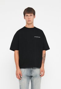 Zwarte katoenen T-shirt met een relaxed fit, met korte mouwen en een witte print waarop "VERSPIL GEEN CULTUUR" staat op de borst.