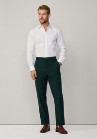 Camicia bianca a maniche lunghe con chiusura a bottoni, abbinata a pantaloni verdi scuri e scarpe in pelle marrone. Design semplice, vestibilità su misura.