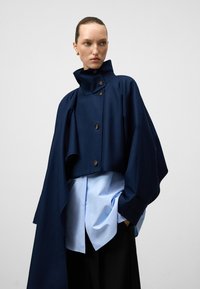 Giacca cropped blu navy con colletto alto e bottoni frontali, indossata sopra una camicia azzurro chiaro con bottoni, caratterizzata da una vestibilità ampia e una texture liscia.
