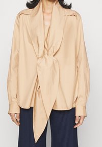 Une femme portant un blouse beige à manches longues avec un grand nœud en tissu à l'avant, associée à un pantalon côtelé marine.