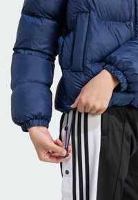 Marine blauwe pufferjack met elastische manchetten en zipzakken, wordt aangepast aan de zoom. Zwart-wit gestreepte sportbroek met drukknopen.