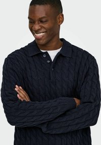 Marinefarbener gestrickter Pullover mit einem Zopfmuster, einem gerippten Polokragen und einem Drei-Knopf-Verschluss. Lange Ärmel mit strukturierten Bündchen.