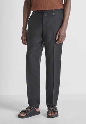 Homme portant un pantalon gris foncé sur mesure, un haut tricoté marron à manches courtes, et des sandales noires à bouts ouverts avec deux brides.