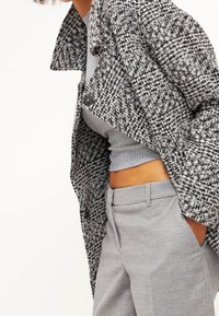 Kabát s šedobílým vzorem houndstooth a strukturovaným límcem, kombinovaný se přiléhavým šedým topem a dobře střiženými šedými kalhotami, které vytvářejí moderní siluetu.