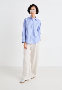 Chemise à rayures bleu clair avec boutons et manches longues, associée à un pantalon en lin beige ample et des baskets bleues, sur un fond blanc.
