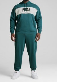 Sudadera verde azulado con franjas blancas y logo, combinada con joggers a juego. Ambos hechos de un tejido suave, con puños acanalados y capucha ajustable.