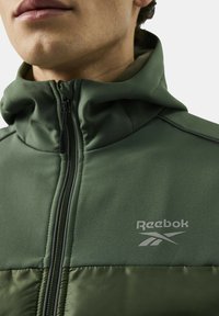 Zielona kurtka na zamek z gładkiej górnej tkaniny i wyłożonym poduszką dolnym sekcją. Na wysokości klatki piersiowej umieszczono logo Reebok w jasnoszarym kolorze.