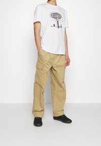 Bílý bavlněný tričko s grafickým potiskem, spárované se béžovými cargo kalhotami s bočními kapsami; černé boty dotvářejí celý outfit.