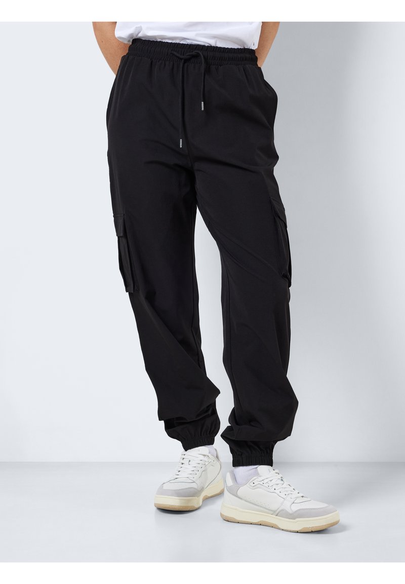 Pantalon cargo noir avec une ceinture élastique, des poches latérales et des chevilles fuselées. Confectionné dans un tissu lisse, présenté avec des baskets décontractées.