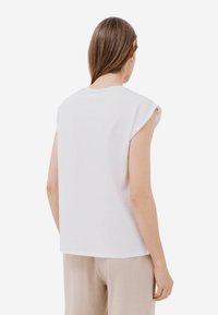 Top bianco senza maniche con scollatura rotonda, texture liscia e cuciture delle spalle arrotolate, abbinato a pantaloni beige chiaro.